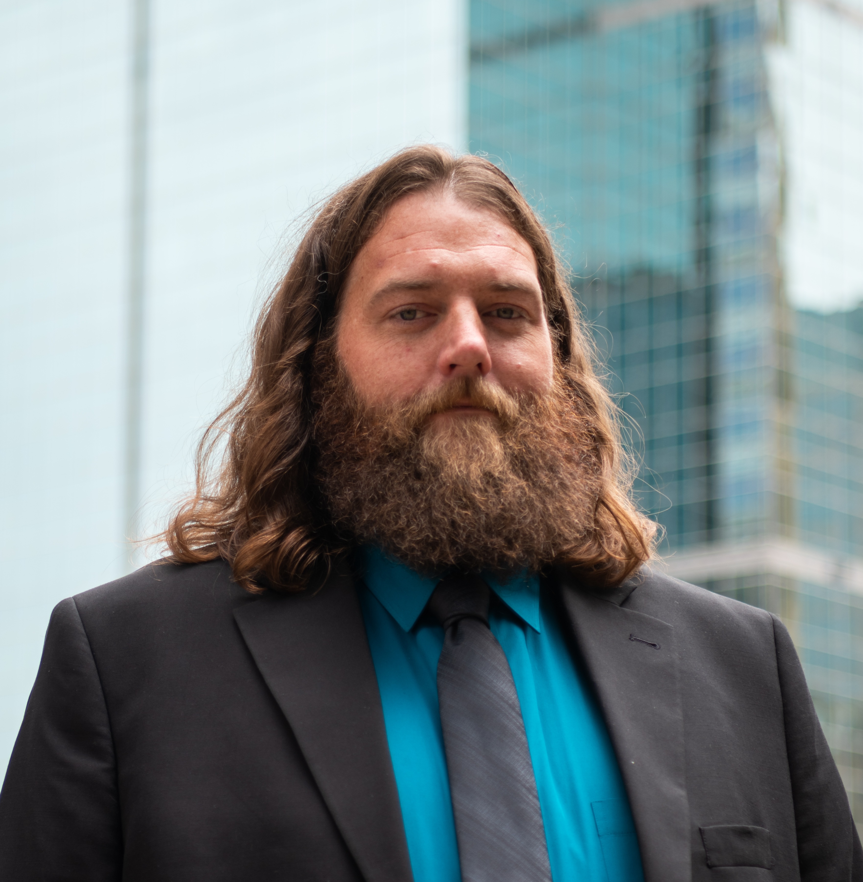 A man with long hair and a beard, wearing a black suit and a teal shirt, stands in front of modern glass buildings.